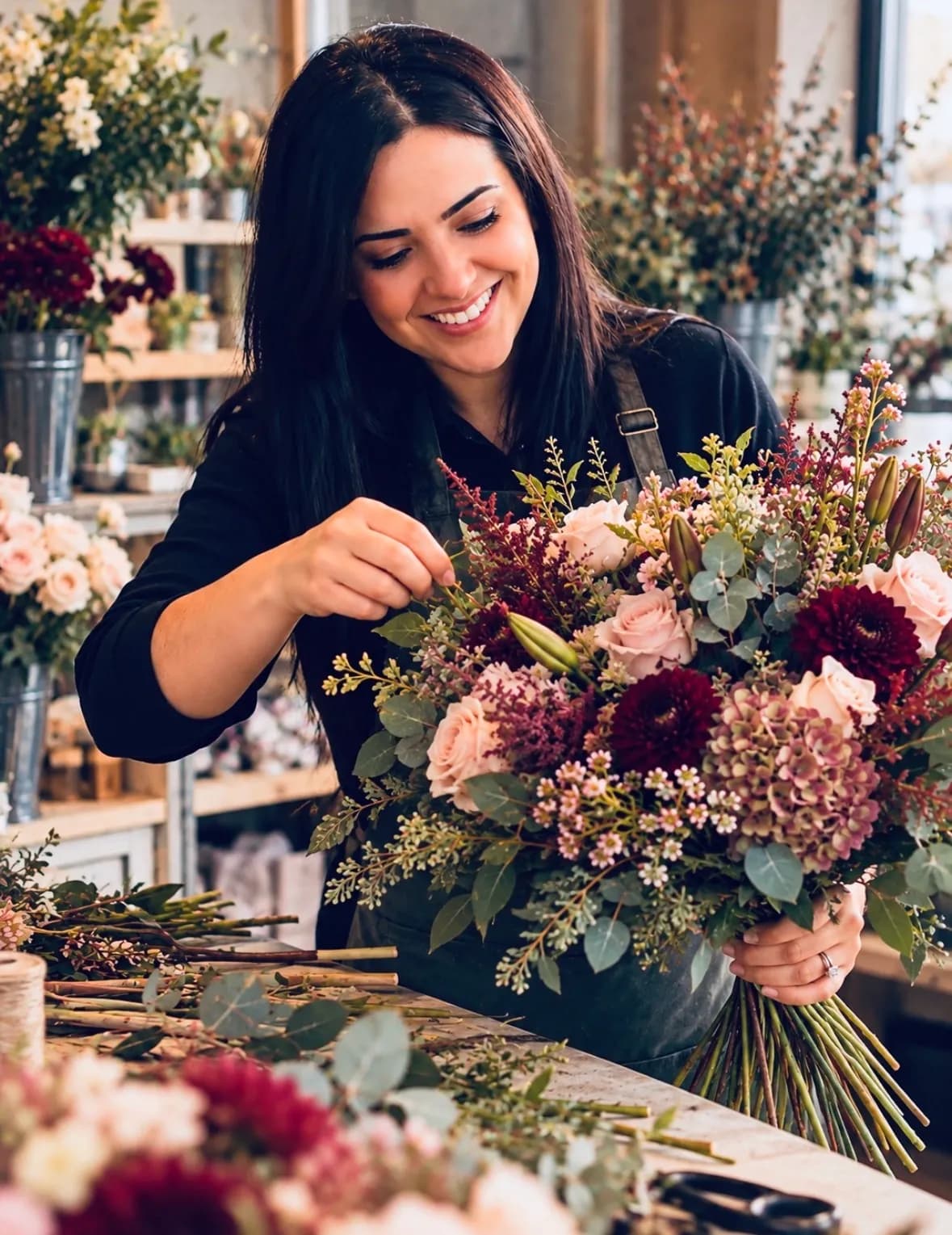 A smiling florist in an apron arranging a large bouquet of pink, red, and white flowers with green foliage, with other flowers and supplies visible in the background.